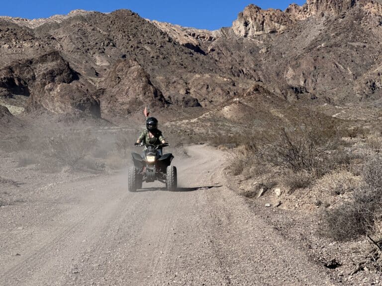Helmeted rider on an ATV explores a dusty Nevada desert trail near Las Vegas with mountain views and blue skies.