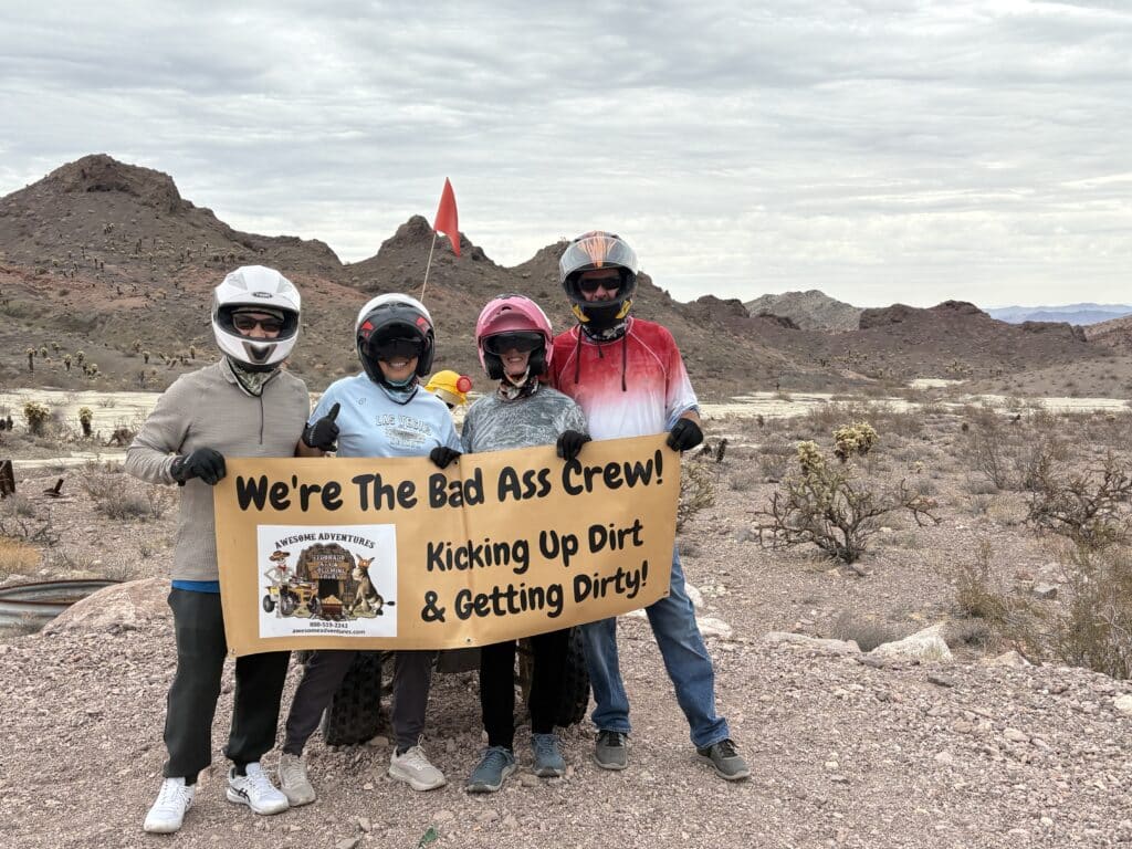 Four adventurers in helmets pose by the Nevada desert with an ATV tour sign, ready for a Las Vegas off-road RZR ride adventure.