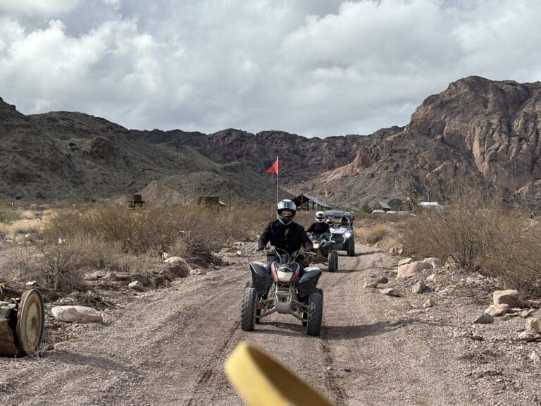 ATV riders explore a Nevada desert trail near Las Vegas, with red flags waving, mountains and dry bushes under cloudy skies.