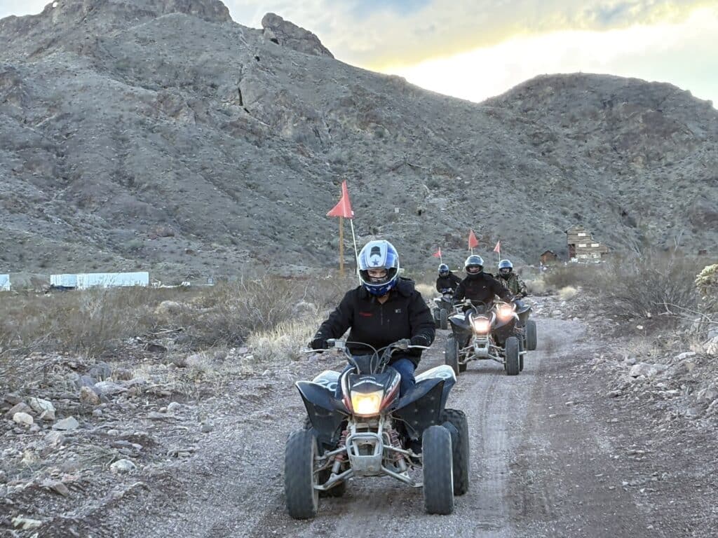 ATV riders explore a rocky Nevada desert trail near Las Vegas, red flags flying, with mountains and Ghost Town sightseeing in view.