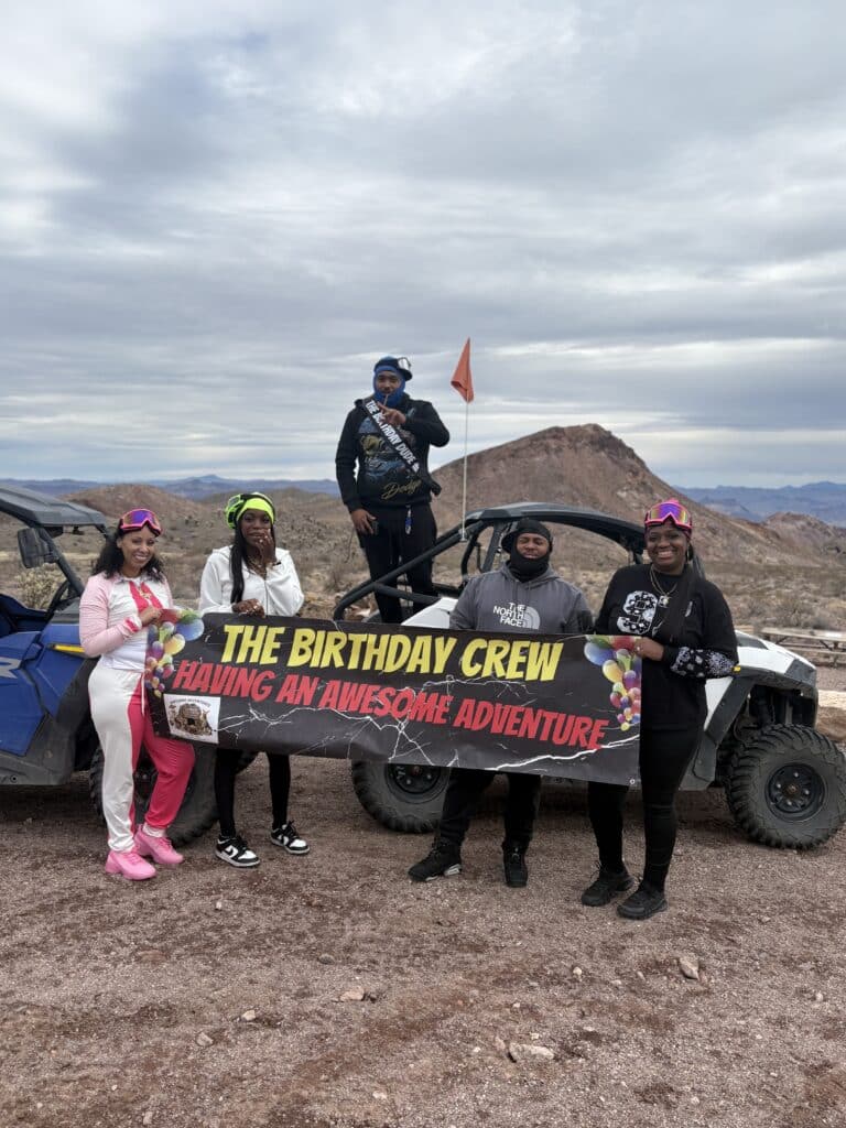 Five friends pose by ATVs in Nevada desert near Las Vegas, holding a “Birthday Crew” banner; RZR tour adventure vibes.