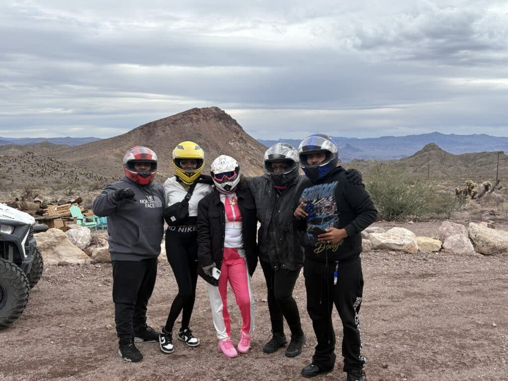 Group in helmets and gear poses on a Nevada desert trail near mountains; ATV vehicle visible—Las Vegas ATV tour adventure.