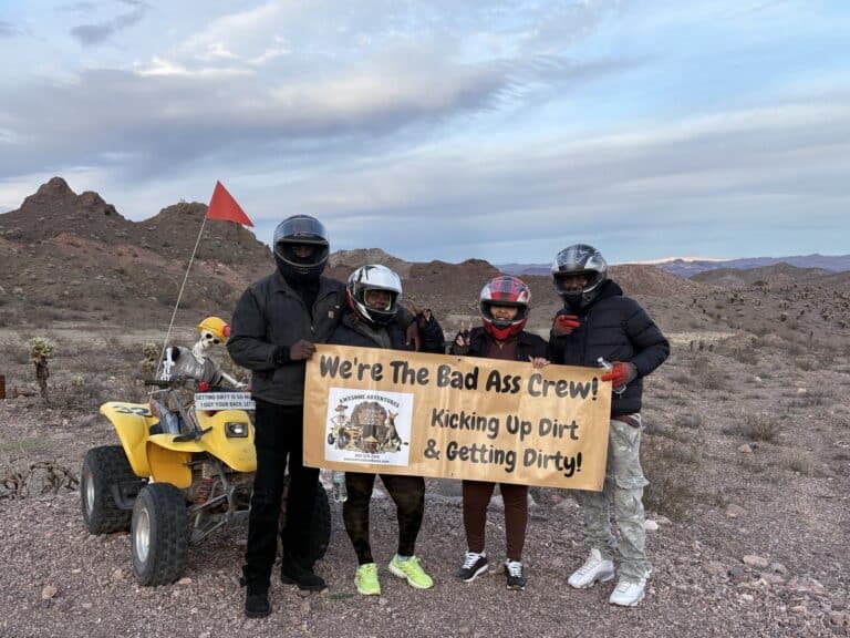 Group in helmets and gear with "Bad Ass Crew" banner by a yellow ATV on a Nevada desert RZR tour near Las Vegas.