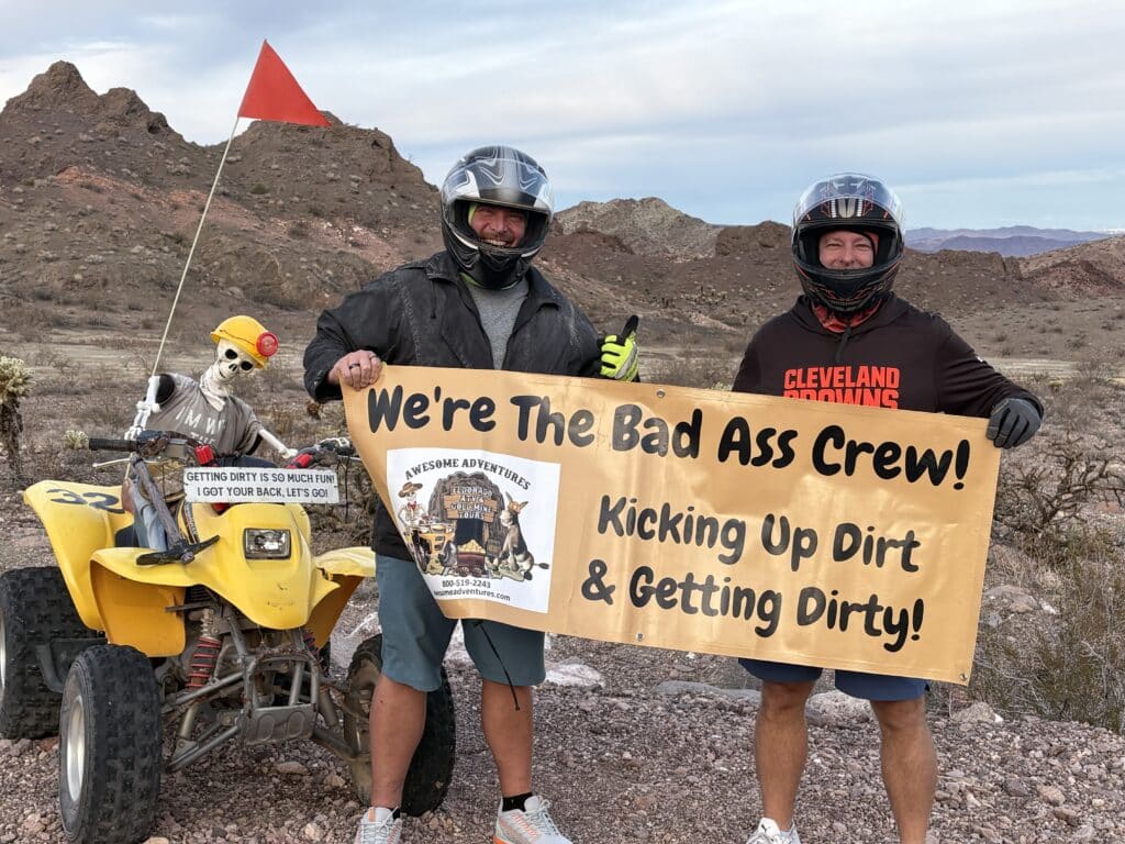 Two men with helmets hold a banner beside a skeleton on an ATV in the Nevada desert near Las Vegas and the Colorado River.