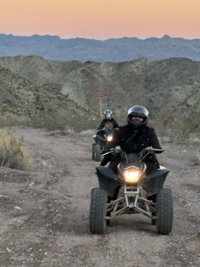 Helmeted riders explore a Nevada desert trail on ATVs at sunset, with the Colorado River and rugged mountains near Las Vegas.