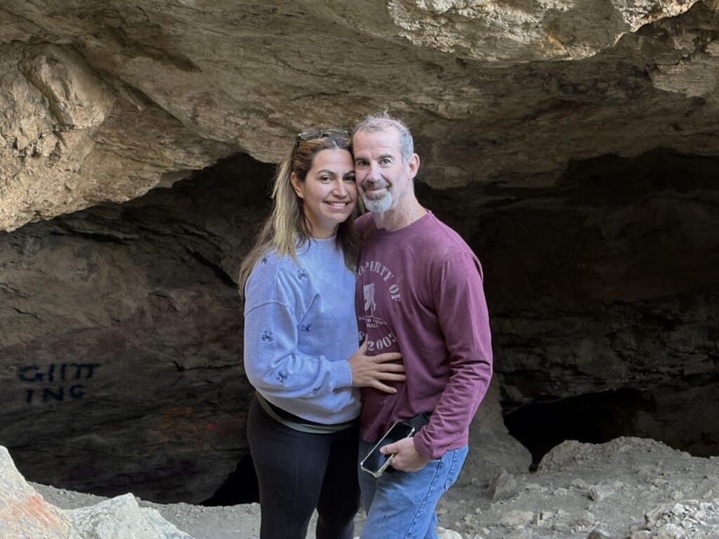 Smiling couple at Nevada cave entrance near Colorado River, dressed for adventure; sunlight highlights rocky walls.