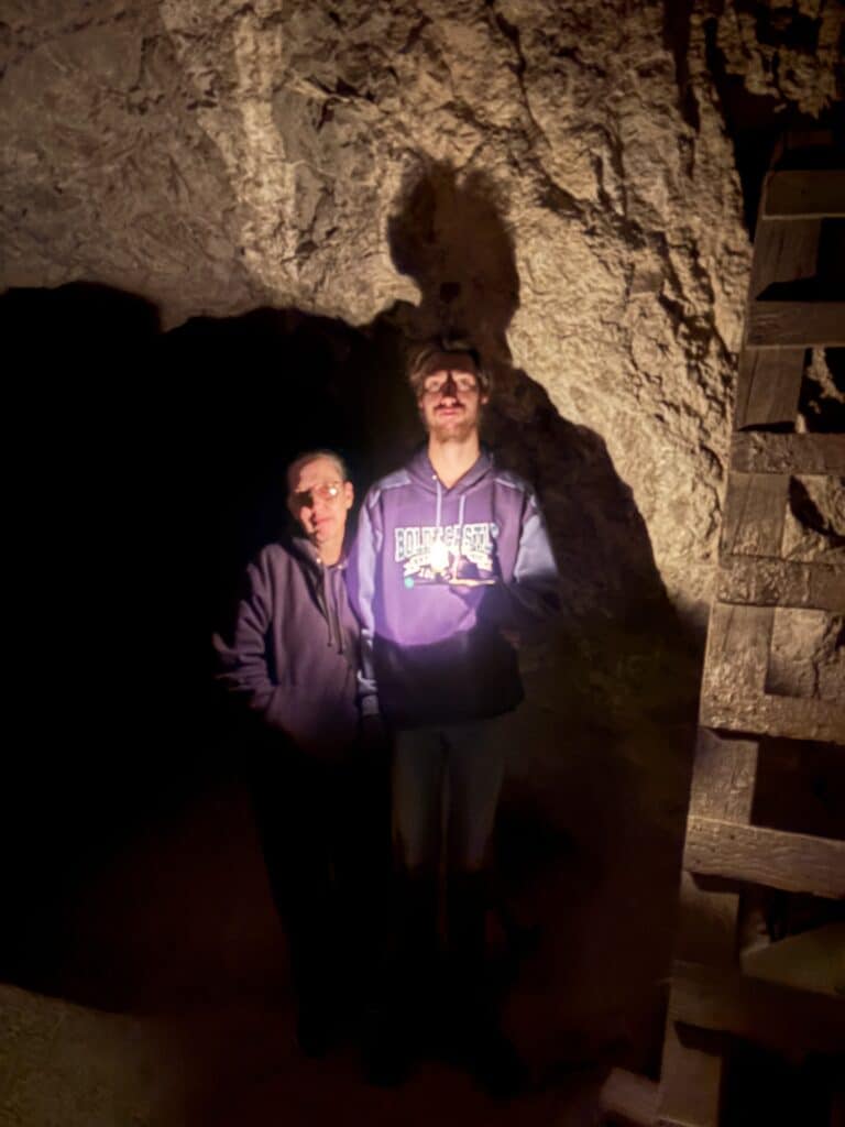 Two people explore a dim cave near a wooden structure on a Las Vegas ATV tour, with dramatic shadows on the Nevada rocks.