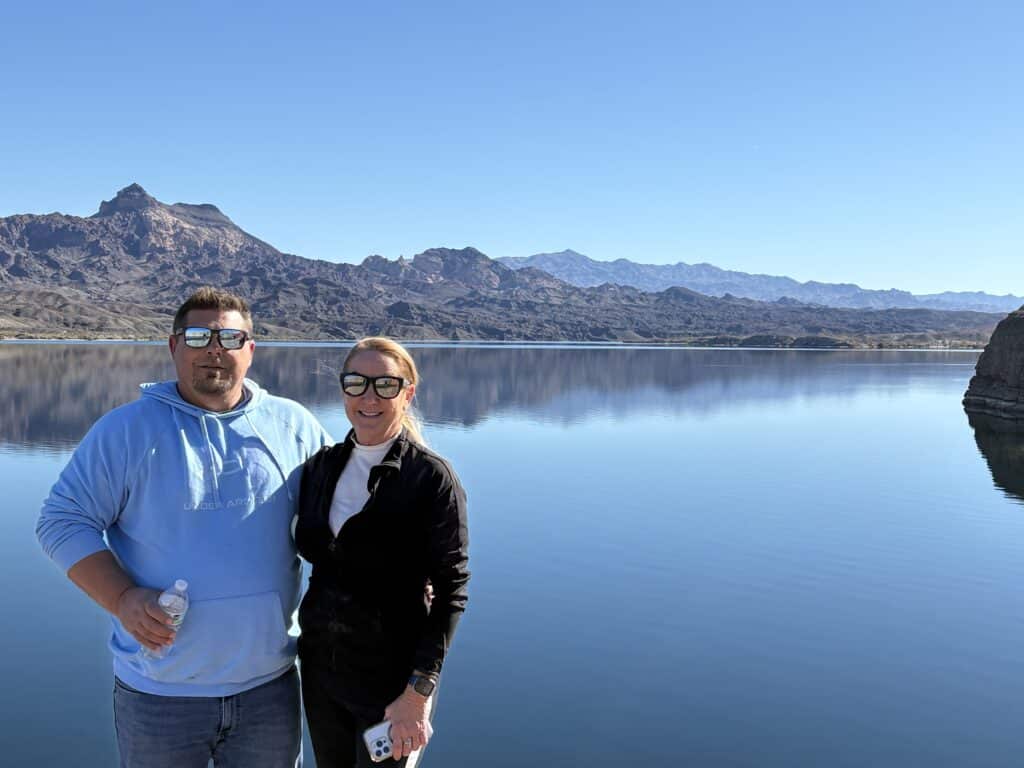Couple smiles by calm Colorado River near Las Vegas, Nevada, with mountains behind. Perfect day for RZR rides or ghost town tours.