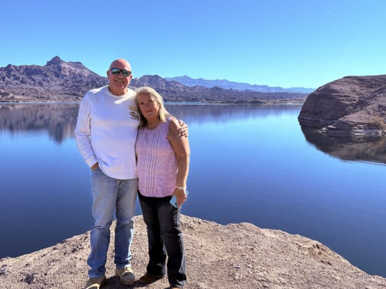 Smiling older couple on rocky Nevada shore by Colorado River near Las Vegas, mountains and clear sky in background.