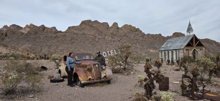 Two people by a vintage car in a Nevada desert ghost town near Las Vegas—old motel, church, cacti; perfect for sightseeing tours.
