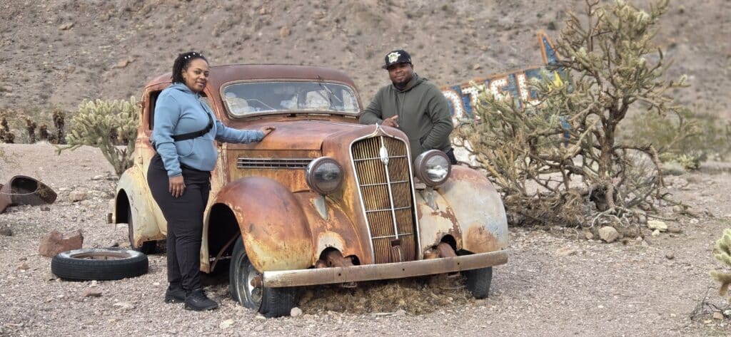 Two people smile by a rusty vintage car in Nevada desert near cacti and rocky hills, perfect for Ghost Town sightseeing.