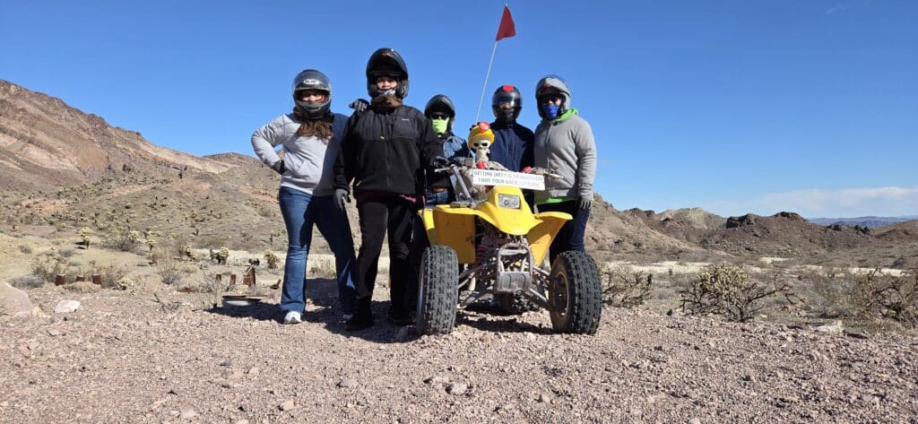 Group in helmets poses by a yellow ATV and skeleton prop in Nevada desert near Las Vegas, ready for ATV tour adventure.