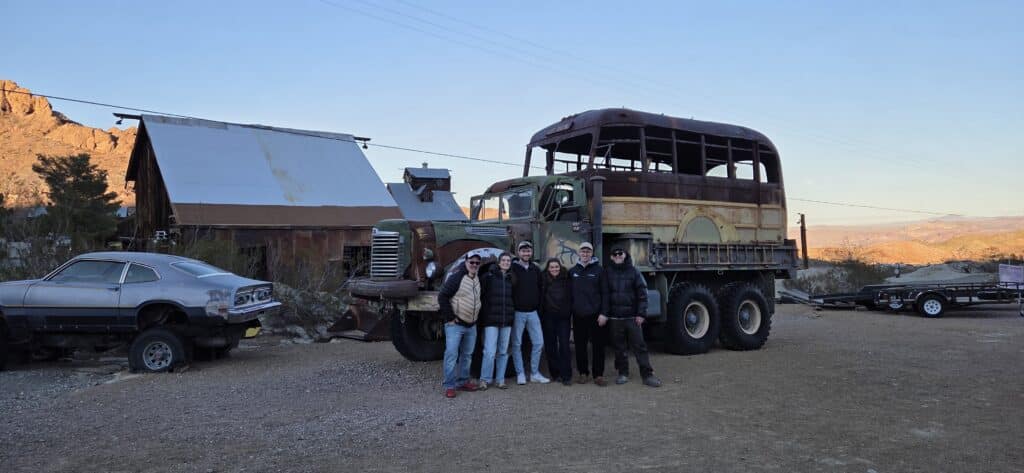 Group posing by vintage double-decker bus near Nevada barn, mountains & car—perfect spot for Las Vegas Ghost Town sightseeing.