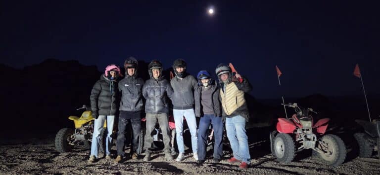 Group in helmets & jackets smiles by ATVs under a moonlit Nevada sky—Las Vegas RZR tour adventure near the Colorado River.