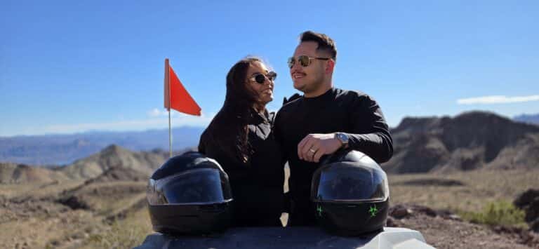 A smiling couple in black by helmets and a red flag during a Nevada ATV tour near the Colorado River and Las Vegas mountains.