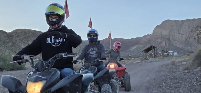 Three helmeted riders on ATVs with red flags explore a Nevada desert trail near Las Vegas as the sun sets, perfect for ATV tours.