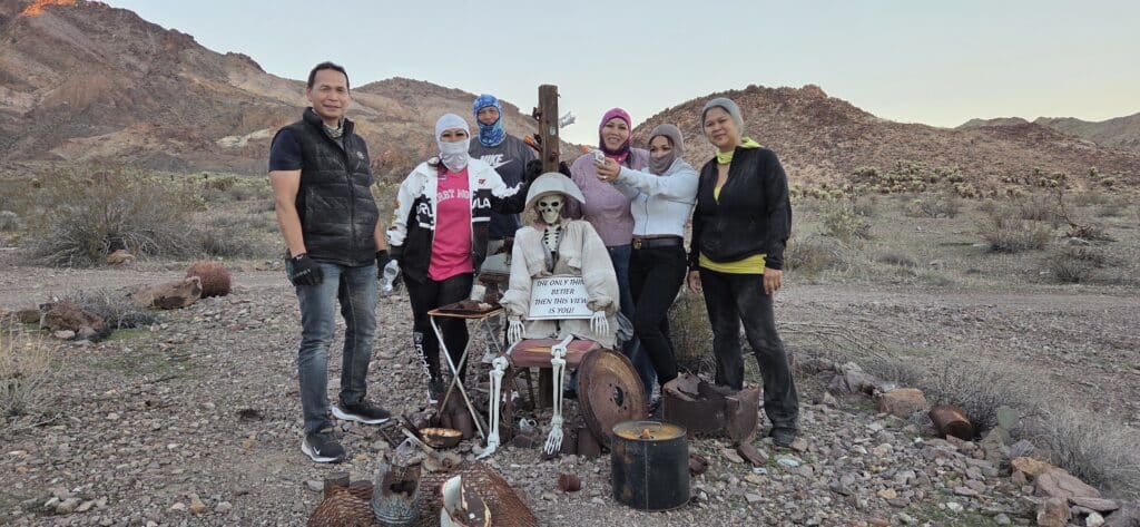 Six friends pose with a skeleton prop near Las Vegas in the Nevada desert, with mountains and cacti, enjoying Ghost Town sightseeing.
