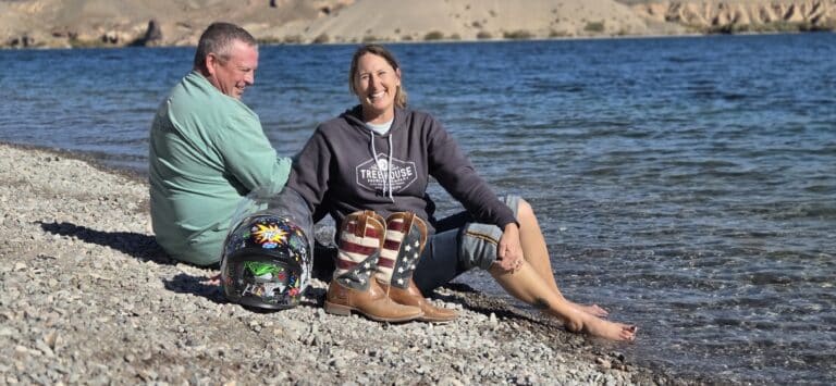 Couple enjoys Colorado River lakeshore near Las Vegas, ATV helmets by side, scenic Nevada hills and water views in the background.