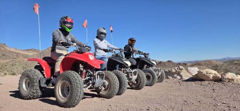 Riders in helmets enjoy an ATV tour on Nevada desert trails near Las Vegas, with rocky hills and orange safety flags in view.