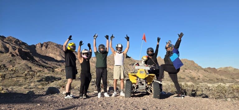 Group in helmets celebrates by yellow ATV on Nevada desert tour near Las Vegas mountains under clear blue sky.