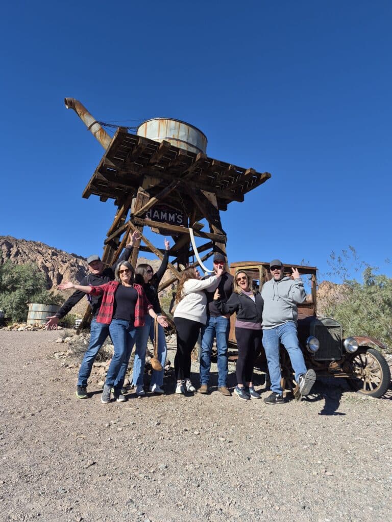 Seven friends pose by a rustic HAMMS water tower and vintage car in a Nevada desert ghost town near Las Vegas under clear skies.