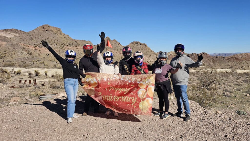 Group celebrating with “Happy Anniversary” banner on Nevada rocky terrain during Las Vegas RZR off-road tour, blue sky behind.