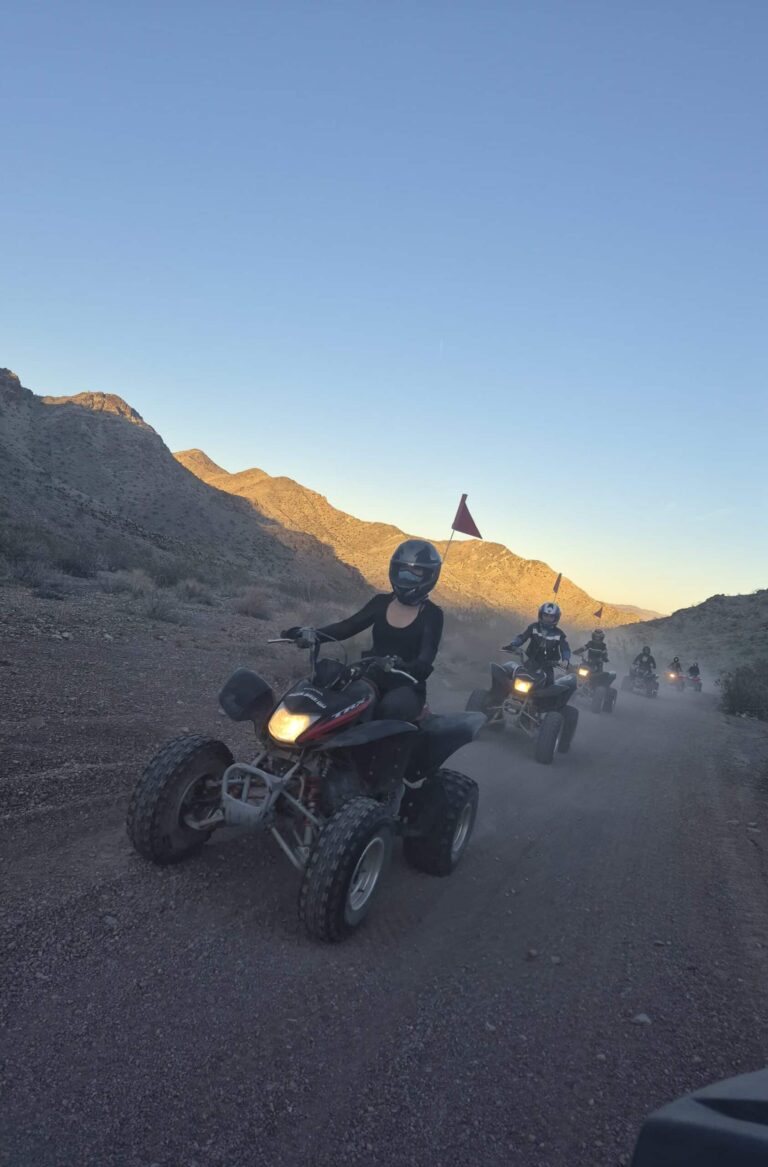 Riders on ATVs explore a dusty Nevada desert trail near Las Vegas at sunset, wearing helmets and red flags during an off-road tour.