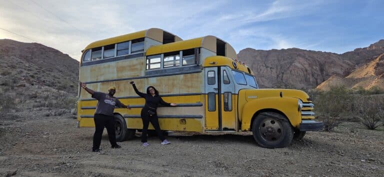 Two people pose with arms outstretched by a yellow school bus near the Colorado River and Nevada mountains, ready for ATV tours.