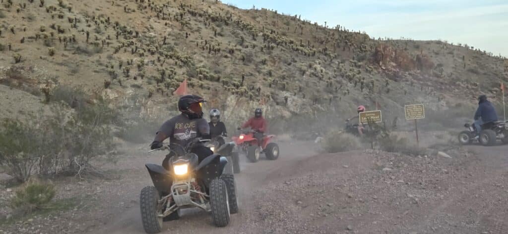 ATV riders explore a dusty Nevada desert trail near Las Vegas, with rocky hills, sparse plants, and yellow trail signs visible.