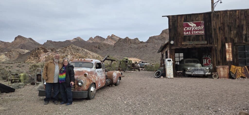 Smiling older couple by vintage car at a Nevada ghost town near Las Vegas, with antique vehicles and desert mountains.