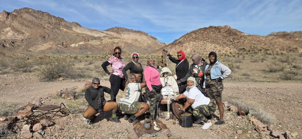 Nine friends smile by a decorated skeleton sign at a Nevada desert camp near rocky hills—perfect for ATV tours or Ghost Town visits.
