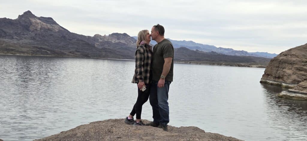 A couple kisses on a rocky Nevada shore by the Colorado River near Las Vegas, with mountains and a cloudy sky in the background.