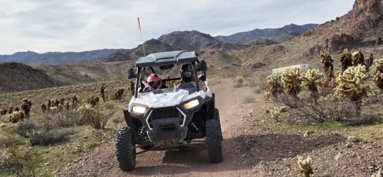 Two people on an ATV tour ride through Nevada's rocky desert near Las Vegas, with cacti, mountains, and dirt trails in view.