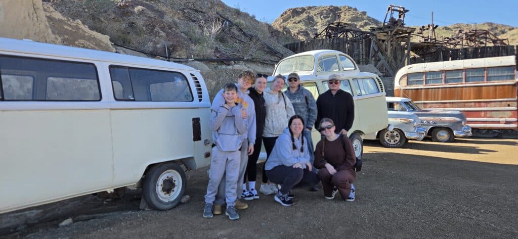 Seven friends pose by a white van near old cars in a Nevada ghost town, with desert mountains and blue sky—perfect for ATV and RZR tours.