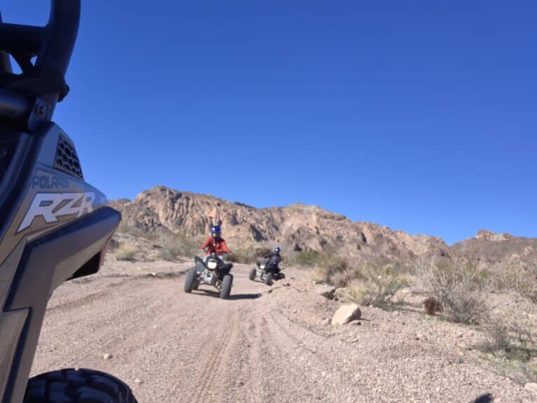 ATV riders explore a dusty Nevada desert trail near Las Vegas, with rocky terrain, mountains, and sparse vegetation in the background.
