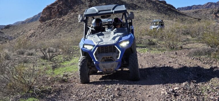 Las Vegas ATV tour: Blue RZR with helmeted riders on rocky desert trail near Nevada mountains, another vehicle behind.