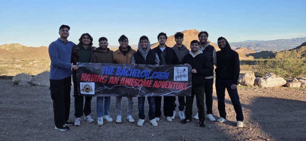 Ten men smile in Nevada desert by rocky hills, holding “The Bachelor Crew” banner after an ATV tour near the Colorado River.