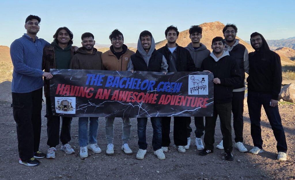Ten men smile in Nevada desert by rocky hills, holding “The Bachelor Crew” banner after an ATV tour near the Colorado River.