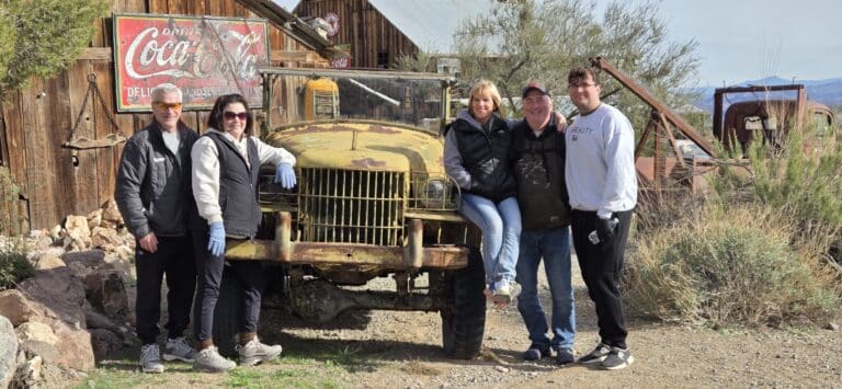 Group poses by vintage yellow truck at Nevada ghost town near Las Vegas, with rustic building and Coca-Cola sign—perfect for sightseeing.