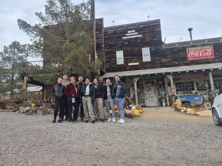 Eight friends smile at a rustic Nevada ghost town near Las Vegas, surrounded by pumpkins and trees on a gravel path.