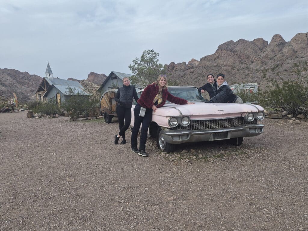 Group poses playfully by a vintage pink car in a Nevada desert ghost town near Las Vegas, with rocky hills and old buildings.