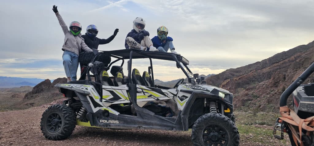 Adventurers in helmets pose on a Polaris RZR during an ATV tour near Las Vegas, Nevada, with mountains and clouds behind.