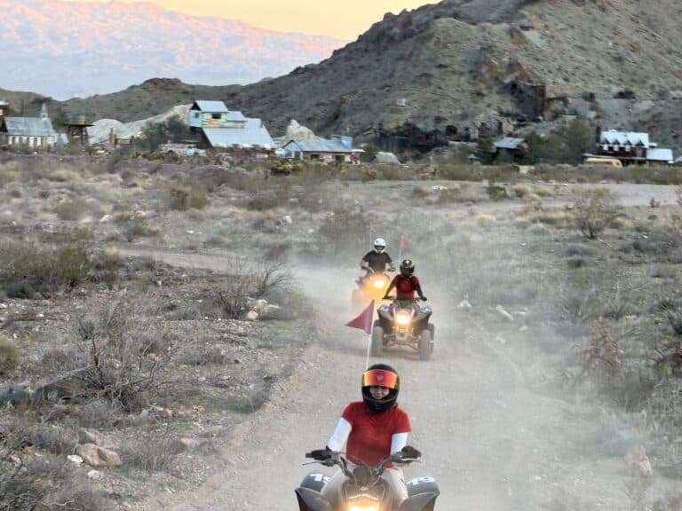 ATV tour near Las Vegas: three riders explore Nevada’s dusty desert, rocky hills, and ghost town buildings at sunset.