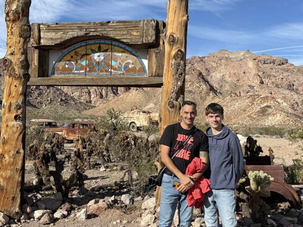 Smiling couple by rustic archway with stained glass in a Nevada ghost town near Las Vegas, cacti, old cars, and desert mountains.