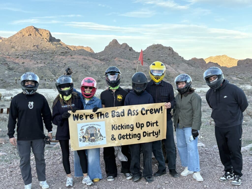 Seven people in helmets hold a “Bad Ass Crew” banner on a Nevada desert ATV tour near Las Vegas and the Colorado River.
