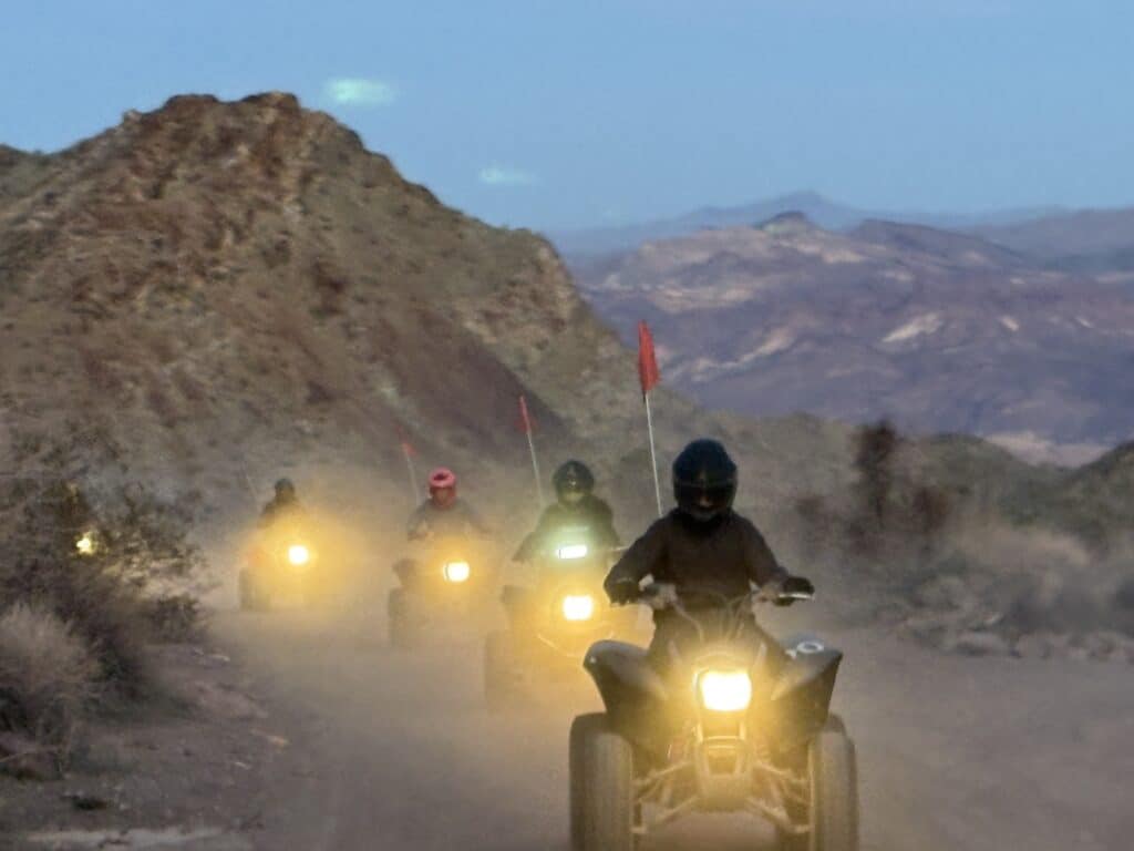 Riders on an ATV tour explore a dusty Nevada trail near Las Vegas at dusk, headlights on and red flags waving in the mountain air.