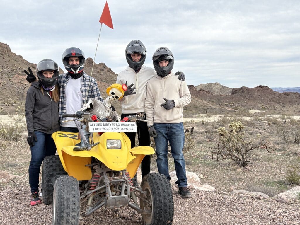 Group in helmets beside yellow ATV with skeleton near Las Vegas, Nevada. Fun ATV tour, peace signs, red flag flying.