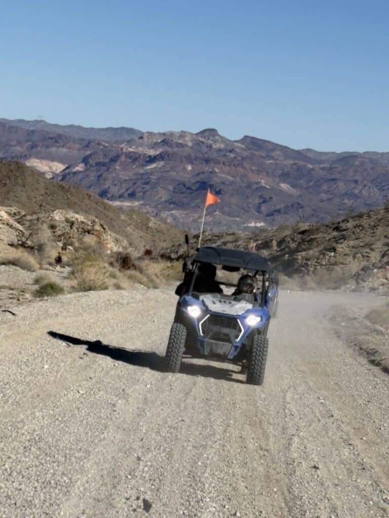 A blue RZR ATV rides a rocky Nevada desert trail near Las Vegas, orange flag flying, mountains and clear sky behind.