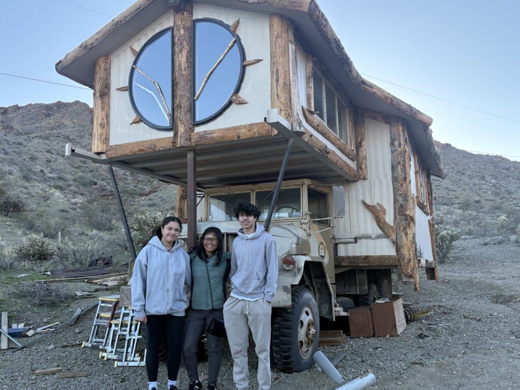 Smiling trio by a wood-paneled tiny house on a military truck in Nevada desert, near Las Vegas ghost towns and ATV tours.