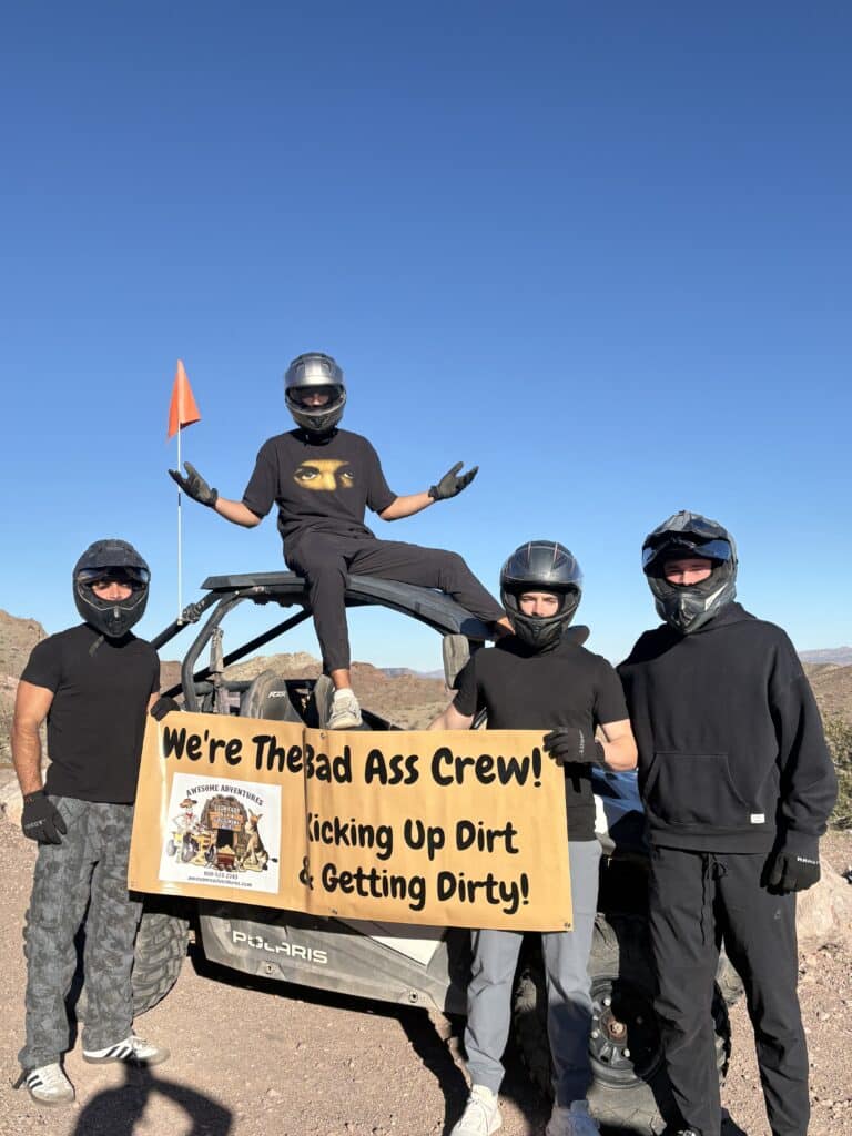 Group in black shirts and helmets by an ATV in Nevada desert, holding a "Bad Ass Crew" sign on Las Vegas off-road RZR tour.
